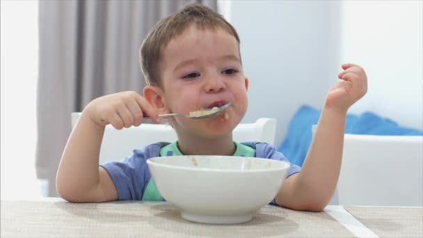 Kis aranyos gyermek ül egy asztalhoz, és enni a saját zabpehely, a baba eszik készségesen. Koncepció boldog gyermekkor. — Stock videók