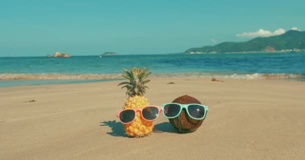 Na tropikalnej plaży zbliżenie owoców w okulary przeciwsłoneczne pod gorącym letnim słońcem wzdłuż tropikalnego egzotycznego wybrzeża, ananasa i kokosowego w okulary przeciwsłoneczne na tle oceanu. Concept lato, Impreza, święta — Wideo stockowe