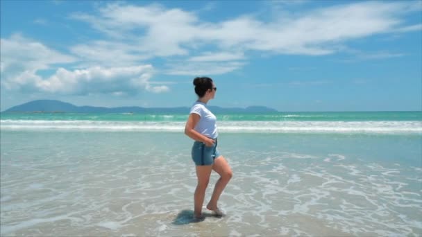 Європейська красива брюнетка в чорних окулярах, біла сорочка і сині шорти йде під спекотним літнім сонцем уздовж тропічного екзотичного узбережжя В'єтнаму, безтурботна дівчина ходить по піску прямо до океану — стокове відео