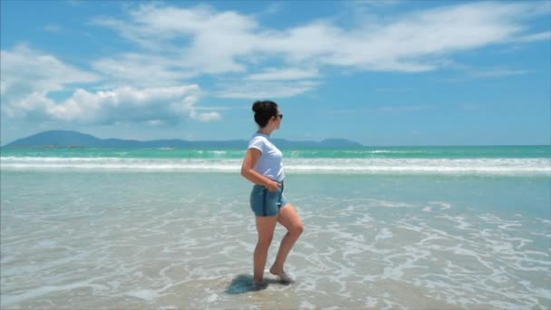 Europäische schöne Brünette in schwarzer Brille, weißem Hemd und blauer Shorts geht unter der heißen Sommersonne entlang der tropischen exotischen Küste Vietnams, unbeschwertes Mädchen, das auf dem Sand bis zum Ozean läuft — Stockvideo