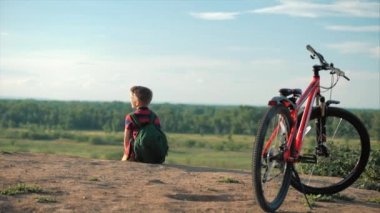 Onun sırt üzerinde bir sırt çantası Ile kırmızı T-shirt Genç, Sunset, yüksek bir tepede oturan, doğa zevk, özgürlük, onun bisiklet arka plan karşı.