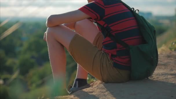 Tonåring i en röd skjorta med en ryggsäck på ryggen, vid solnedgången, sitter på en hög kulle och tittar på molnen i himlen, natur, flod, träd. — Stockvideo
