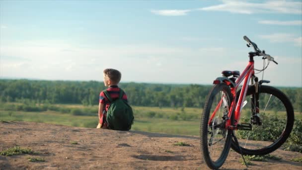 Tonåring i en röd T-shirt med en ryggsäck på ryggen, vid solnedgången, sittande på en hög kulle, njuter av naturen, frihet, mot bakgrund av sin cykel. — Stockvideo