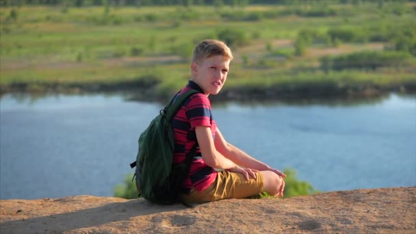 Nahaufnahme Porträt kaukasischer Teenager in rotem Hemd mit Rucksack auf dem Rücken, bei Sonnenuntergang auf einem hohen Hügel sitzend, den warmen Sommerurlaub genießend. — Stockvideo