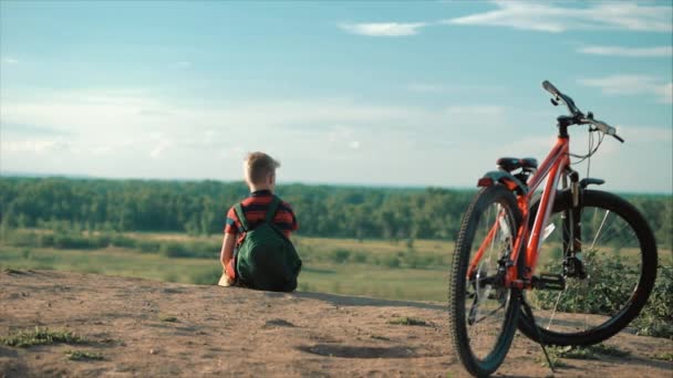 Tonåring i en röd T-shirt med en ryggsäck på ryggen, vid solnedgången, sittande på en hög kulle, njuter av naturen, frihet, mot bakgrund av sin cykel. — Stockvideo