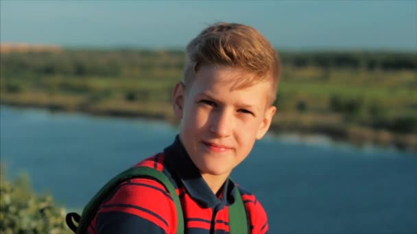 Close up portret Kaukasische jongen tiener in een rood shirt met een rugzak op zijn rug, bij zonsondergang, zittend op een hoge heuvel genieten van warme zomervakantie. — Stockvideo