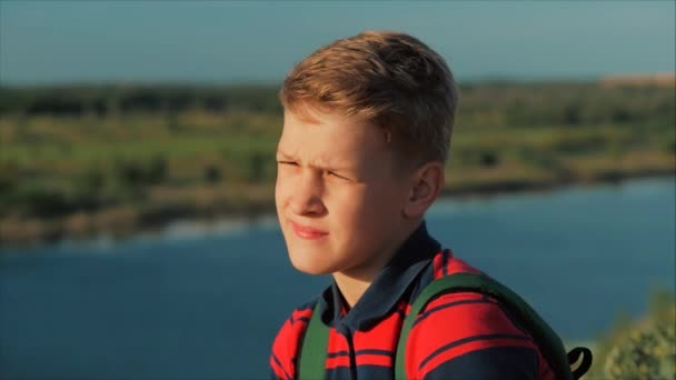 Adolescente con una camisa roja con una mochila en la espalda, al atardecer, sentado en una colina alta y mirando las nubes en el cielo, la naturaleza, el río, los árboles . — Vídeos de Stock