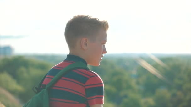 Adolescente em uma camisa vermelha com uma mochila nas costas, ao pôr do sol, sentado em uma colina alta e olhando para as nuvens no céu, natureza, rio, árvores . — Vídeo de Stock