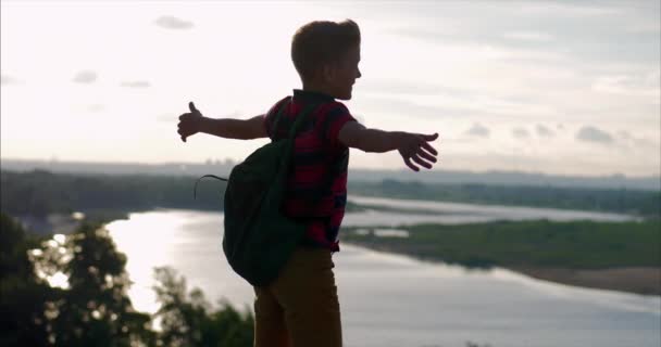 Vid Sunset Child i en röd skjorta en ryggsäck på ryggen, nå till toppen på en hög kulle och titta på molnen i himlen, natur, stående på toppen av ett berg, händer upp, medvetenhet om framgång — Stockvideo