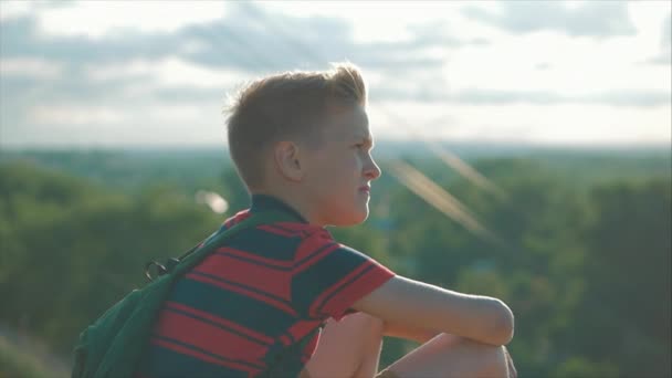 Tonåring i en röd skjorta med en ryggsäck på ryggen, vid solnedgången, sitter på en hög kulle och tittar på molnen i himlen, natur, flod, träd. — Stockvideo