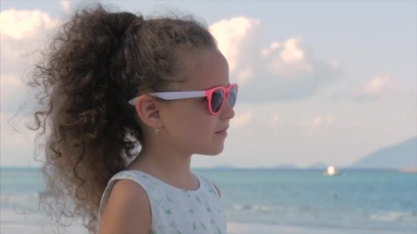 Close-up portret van een mooi klein meisje in roze bril, schattig glimlachen, kijkend naar de zee. Concept: kinderen, kinderjaren, zomer. — Stockvideo