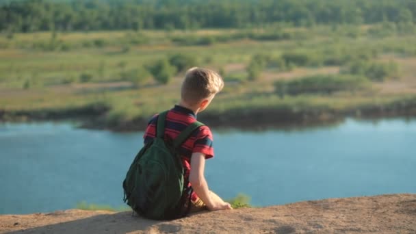 Teenager in a red shirt with a backpack on his back, at sunset, sitting on a high hill and looking at the clouds in the sky, nature, river, trees. — Stock Video