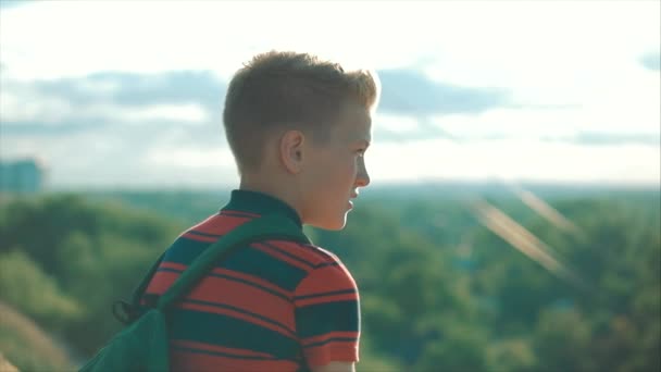 Tiener in een rood shirt met een rugzak op zijn rug, bij zonsondergang, zittend op een hoge heuvel en kijkend naar de wolken in de hemel, natuur, rivier, bomen. — Stockvideo