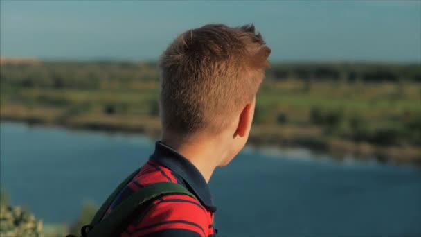 De cerca Retrato Caucásico Niño Adolescente en una camisa roja con una mochila en la espalda, al atardecer, sentado en una colina disfrutando de unas cálidas vacaciones de verano . — Vídeos de Stock