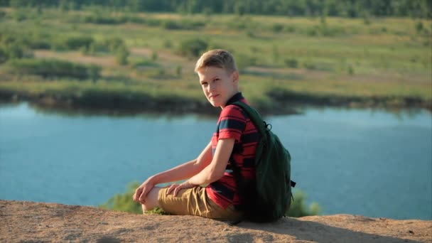 Ein Teenager im roten Hemd mit einem Rucksack im Rücken, bei Sonnenuntergang, auf einem hohen Hügel sitzend, die Natur, die Freiheit genießend, in die Kamera blickend, lächelnd. — Stockvideo
