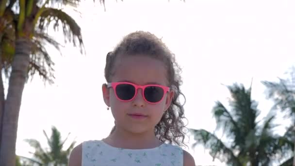 Κοντινό-up πορτραίτο ενός όμορφου μικρού κοριτσιού σε ροζ γυαλιά, χαριτωμένο χαμόγελο, κοιτάζοντας την κάμερα. Έννοια: παιδιά, παιδική ηλικία, καλοκαίρι, μωρό κορίτσι. — Αρχείο Βίντεο