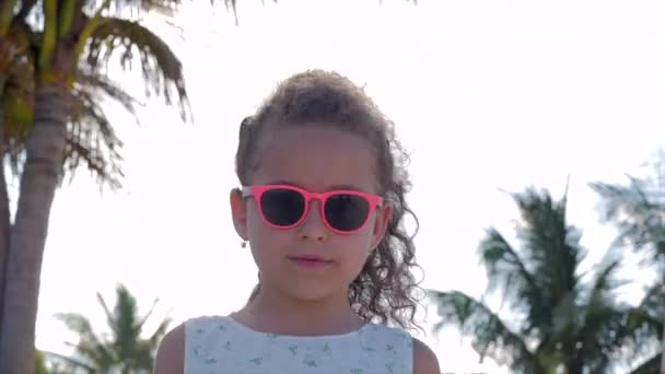 Primer plano Retrato de una hermosa niña en gafas rosadas, linda sonrisa, mirando a la cámara. Concepto: Niños, Infancia, Verano, Niña . — Vídeo de stock