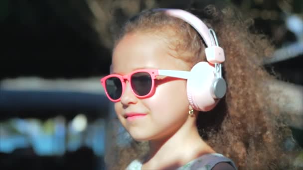 Retrato de un niño lindo, una niña hermosa maravillosa en un vestido blanco con gafas rosadas y auriculares rosados, mirando a la cámara, escuchando música. Concepto Infancia Feliz . — Vídeo de stock