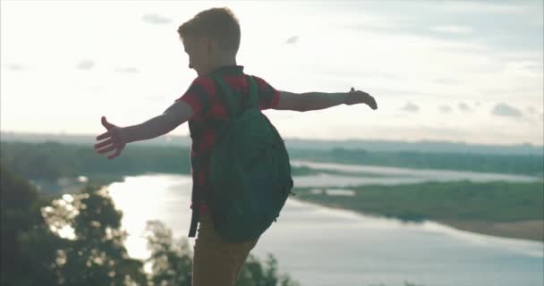 Vid Sunset Child i en röd skjorta en ryggsäck på ryggen, nå till toppen på en hög kulle och titta på molnen i himlen, natur, stående på toppen av ett berg, händer upp, medvetenhet om framgång — Stockvideo