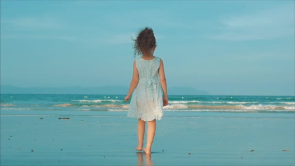 Niña rizada en vestido blanco caminando por la playa al atardecer. A cámara lenta. Concepto de Infancia, Libertad y Viaje Feliz . — Vídeo de stock