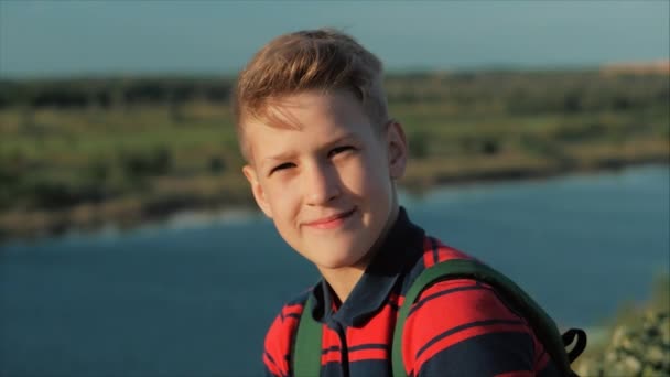 Retrato Joven Caucásico Niño Adolescente en una camisa roja con una mochila en la espalda, al atardecer, sentado en una colina disfrutando de unas cálidas vacaciones de verano . — Vídeos de Stock