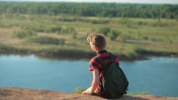 Tonåring i en röd skjorta med en ryggsäck på ryggen, vid solnedgången, sitter på en hög kulle och tittar på molnen i himlen, natur, flod, träd. — Stockvideo