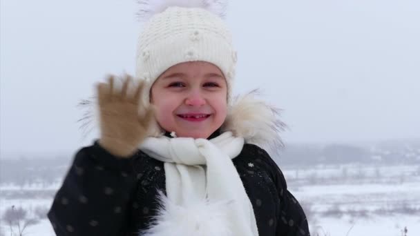 CU Little Girl in a White Knitted Hat Beautiful Smiling Looking at the Camera, it is Snowing on the Street, She looks at the Camera and Waving and Smiling. Концепция детства зимой . — стоковое видео