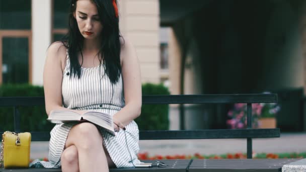 Femme mignonne dans le parc avec une jante rouge sur la tête s'assoit sur un banc et lit un livre . — Video