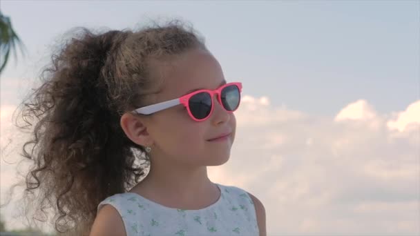 Close-up portret van een mooi klein meisje in roze bril, schattig glimlachen, kijkend naar de blauwe hemel. Concept: kinderen, kindertijd, zomer, baby, peuter. — Stockvideo