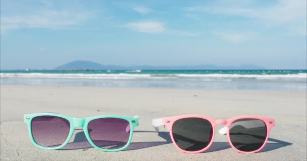 На тропічному пляжі крупним планом сонцезахисні окуляри рожево-сині лежать на білому піску біля океану . — стокове відео