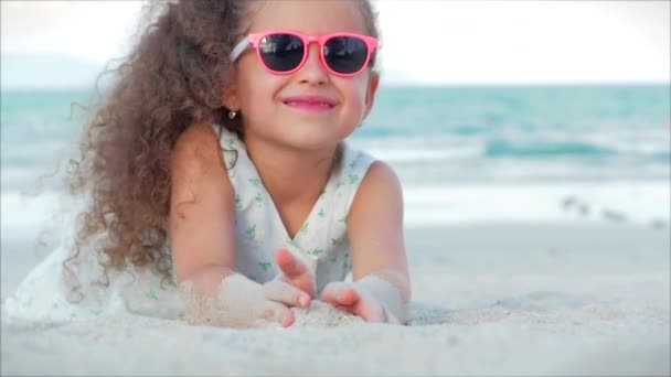Portrét krásné malé holčičky v růžových brýlích, roztomilá usměvavá dívenka na kameru, ležící na písku u moře, nalité písek z ruky. — Stock video