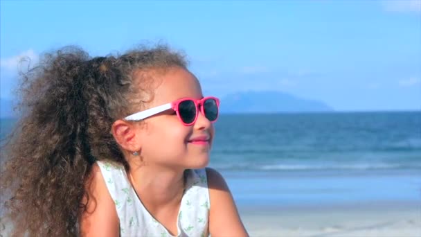 Portret pięknej dziewczynki w różowe okulary, cute uśmiechnięty patrząc na kamerę, leżąc na piasku nad morzem, pours piasek ręki. — Wideo stockowe