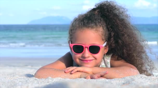 Portret van een mooi klein meisje in roze bril, schattig glimlachend kijkend naar de camera, liggend op het zand aan zee, giet zand van de hand. — Stockvideo