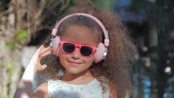 Portrét roztomilýho dítěte, nádherné malé krásné dívky v bílých šatech s růžovými brýlemi a růžovým sluchátky, s pohledem na kameru a naslouchající hudbě. Koncepce šťastné dětství. — Stock video