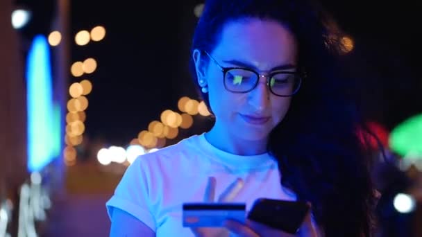 Pretty Woman se encuentra en la calle en una camiseta blanca y gafas, hace pagos en línea con tarjeta de crédito y teléfono inteligente, tecnología de compras en línea. Pago por compra con tarjeta de crédito y teléfono móvil . — Vídeos de Stock