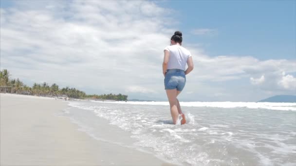 Europese mooie brunette in zwarte glazen, wit-shirt en blauwe broek gaat onder de hete zomer zon langs de tropische exotische kust van Vietnam, zorgeloos meisje lopen op het zand recht op de Oceaan — Stockvideo