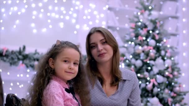 Joven y atractiva madre e hija sentadas abrazadas mirando a la cámara sonriendo sobre el fondo de las luces de Navidad y el árbol de Navidad . — Vídeos de Stock