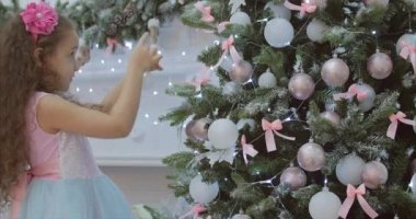 Noel süslemeleri Noel ışıkları ile Noel ağacı üzerinde asılı cu küçük kız şenlikli elbiseli, kıvırcık saçlı. Bir top ile Noel ağacı süs. 4k
