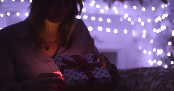 Menina branca bonito sentado no quarto no fundo das luzes de Natal e abrir uma caixa com um presente, sorrindo, segurando-o em suas mãos um presente de desejo . — Vídeo de Stock
