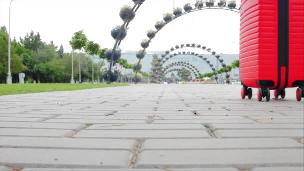 Spor ayakkabılı kadın yürür ve kırmızı bir bavulu tekerlekli olarak yuvarlar. Bir kadın bavuluyla sokakta yürüyor. Seyahat kavramı. Yavaş çekim. — Stok video