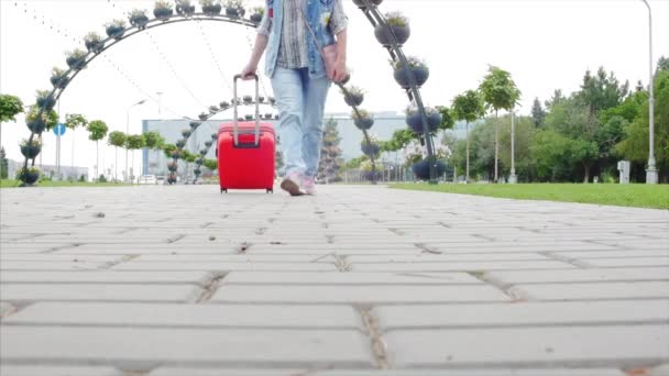 Η γυναίκα στα αθλητικά περπατάει και κάνει μια κόκκινη βαλίτσα με ρόδες. Μια γυναίκα περπατάει στο δρόμο με τη βαλίτσα της. Η έννοια του ταξιδιού. Αργή κίνηση. — Αρχείο Βίντεο