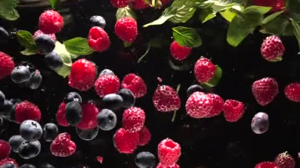Fresh raspberries with blueberries and fresh mint leaves fall on a black background into the water, splashing on a black background. Drop fresh fruits and berries into the water. Healthy eating, slow — Stock Video