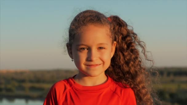 Портрет счастливой маленькой девочки, глядящей в камеру, на закате под открытым небом. След фондового рынка . — стоковое видео