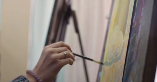 Ung kvinnlig konstnär är i en konst Studio, sitter bakom en staffli och målning på duk. Ritning process: i konsten studion av konstnärerna hand konst flicka med en pensel målning på duk. 4K — Stockvideo