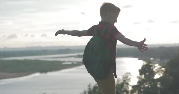 Tonåring i en röd skjorta med en ryggsäck på ryggen, vid solnedgången, sitter på en hög kulle och tittar på molnen i himlen, natur, flod, träd. — Stockvideo