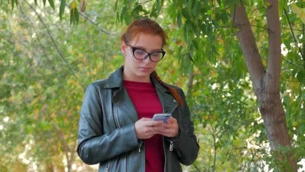 Atractiva joven sonriente con gafas de una mujer feliz con el pelo largo y rojo de buen humor, emociones positivas, escribiendo en un teléfono móvil . — Vídeos de Stock