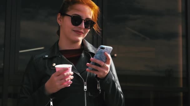 Mujer joven bebiendo café en la calle, escribiendo mensajes de texto en un teléfono móvil, mientras camina en un entorno urbano . — Vídeos de Stock