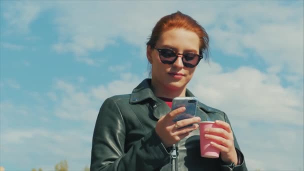 Fiatal nő iszik kávé az utcán, gépelés textil egy mozgatható telefon, rövid idő gyaloglás-ban egy városi elintézés. — Stock videók