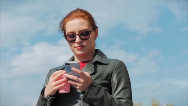 Mujer joven bebiendo café en la calle, escribiendo mensajes de texto en un teléfono móvil, mientras camina en un entorno urbano . — Vídeo de stock