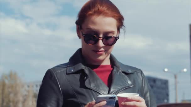 Jovem mulher bebendo café na rua, digitando mensagens de texto de um telefone celular, enquanto caminha em um ambiente urbano . — Vídeo de Stock